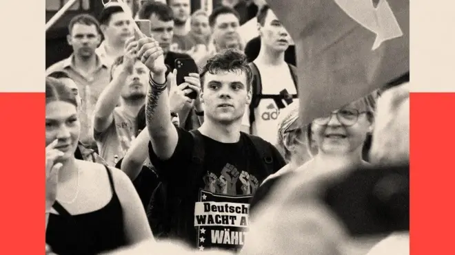 Толпа людей с флагами, в центре кадра молодой мужчина в футболке с изображением сжатых кулаков, он тоже держит флаг.